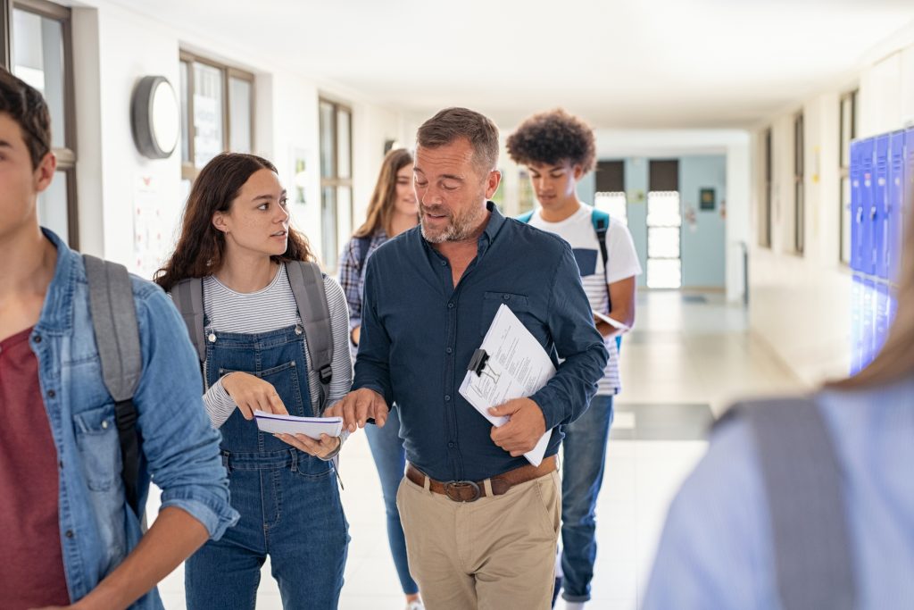 High school student asking doubt to professor while walking in hallway. Mature man professor answering to girl in university campus. High school lecturer talking to schoolgirl at the end of the lesson while walking in the corridor.